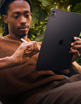 A person outside by foliage holding their iPad Pro in one hand and Apple Pencil Pro in the other